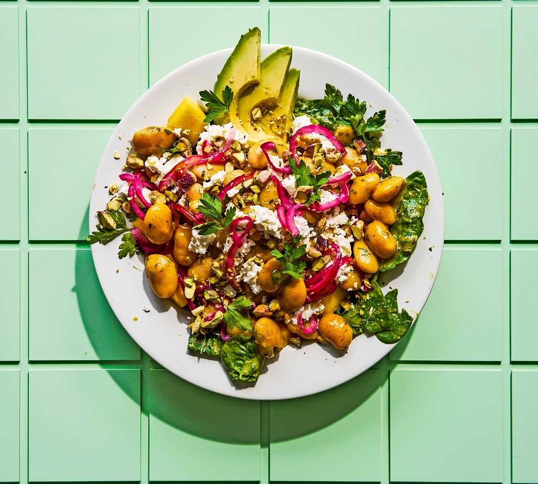 Overhead view of a salad with chickpeas, feta cheese, avocado, red onion, and pistachios on a light green tiled surface.